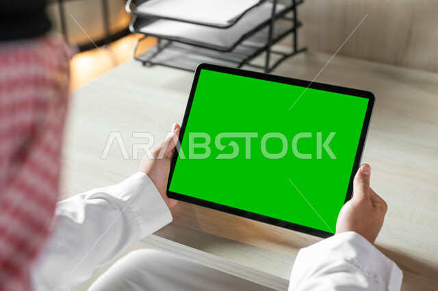 Using a tablet to work remotely, completing work quickly and easily, holding meetings via the Internet, technical and technological development in various fields, a close-up photo from the back of a Saudi Gulf Arab man wearing traditional clothing and a shemagh, holding the tablet in his hand with a blank green screen, chroma