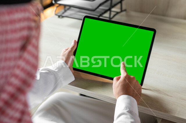 E-learning through the use of modern technologies, a photograph from the back of a Saudi Gulf Arab man holding a portable tablet with a blank green screen in his hand, making video calls and live broadcasts, following favorite movies and series online, Chroma