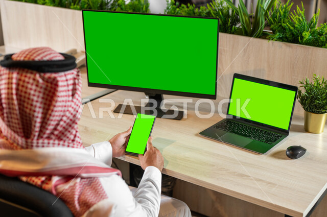 القيام بالدردشات والمحادثات مع العائلة والأصدقاء، إنجاز مهام العمل عبر الإنترنت، استخدام تقنية سمارت على الموبايل للتحكم بشاشة العرض، تصوير مقرب من الخلف لرجل عربي خليجي سعودي يحمل بيده الهاتف المحمول، أجهزة إلكترونية بشاشة خضراء فارغة، كروما