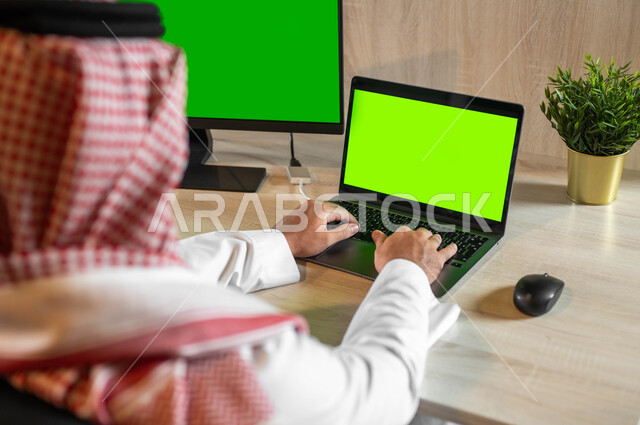 Searching for work via the Internet, technical and technological development in various fields, a picture from the back of a Saudi Gulf Arab man completing practical tasks via a computer, following social media news, electronic devices with a blank green screen, chroma