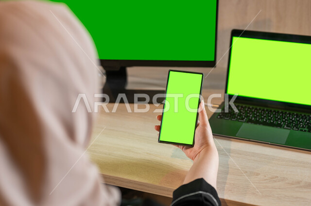 Technical and technological development in several fields, electronic devices with a blank green screen, a photograph from the back of a Saudi Gulf Arab woman sitting in front of a desk with a mobile phone in her hand, the use of modern devices and technologies to facilitate work, Chroma