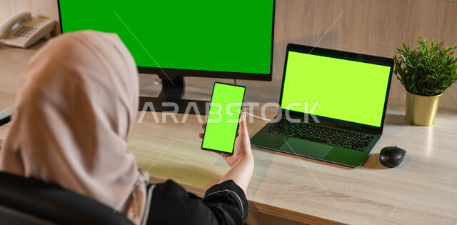 أجهزة الكترونية بشاشة خضراء فارغة ، استخدام الأجهزة والتقنيات الحديثة لتسهيل العمل، تصوير مقرب من الخلف لامرأة عربية خليجية سعودية تجلس أمام مكتب وفي يدها هاتف محمول، التطور التقني والتكنولوجي في عدة مجالات، كروما