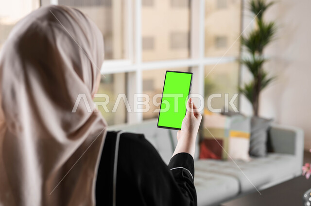 التحكم بالتلفاز من خلال استخدام تقنية سمارت على الهاتف المحمول، متابعة أخبار السوشال ميديا ومواقع التواصل الإجتماعي، تصوير مقرب من الخلف لامرأة عربية خليجية سعودية تحمل بيدها موبايل بشاشة خضراء فارغة، المحادثات والدردشات مع العائلة والأصدقاء، كروما