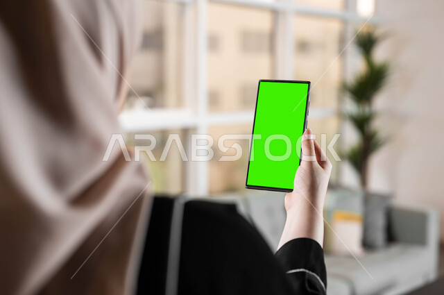 تصفح الانترنت و متابعة أخبار السوشال ميديا ومواقع التواصل الإجتماعي، استخدام تقنية سمارت على الهاتف المحمول للتحكم بالتلفاز، تصوير مقرب من الخلف لامرأة عربية خليجية سعودية تحمل بيدها موبايل بشاشة خضراء فارغة، المحادثات والدردشات مع العائلة والأصدقاء، كروما