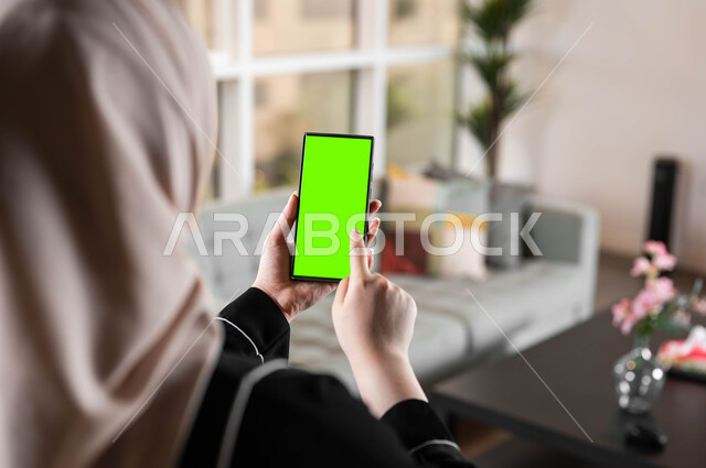 المحادثات والدردشات مع العائلة والأصدقاء، استخدام تقنية سمارت على الهاتف المحمول للتحكم بالتلفاز، تصوير مقرب من الخلف لامرأة عربية خليجية سعودية تحمل بيدها موبايل بشاشة خضراء فارغة، متابعة أخبار السوشال ميديا ومواقع التواصل الإجتماعي، كروما