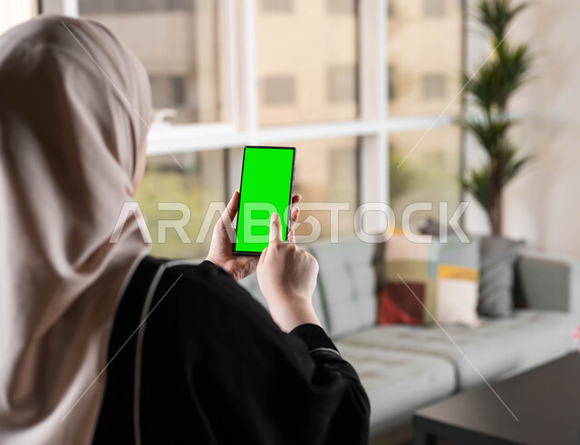 الدردشات والمحادثات مع العائلة والأصدقاء، استخدام تقنية سمارت على الهاتف المحمول للتحكم بالتلفاز، تصوير مقرب من الخلف لامرأة عربية خليجية سعودية تحمل بيدها موبايل بشاشة خضراء فارغة، متابعة أخبار السوشال ميديا ومواقع التواصل الإجتماعي، كروما