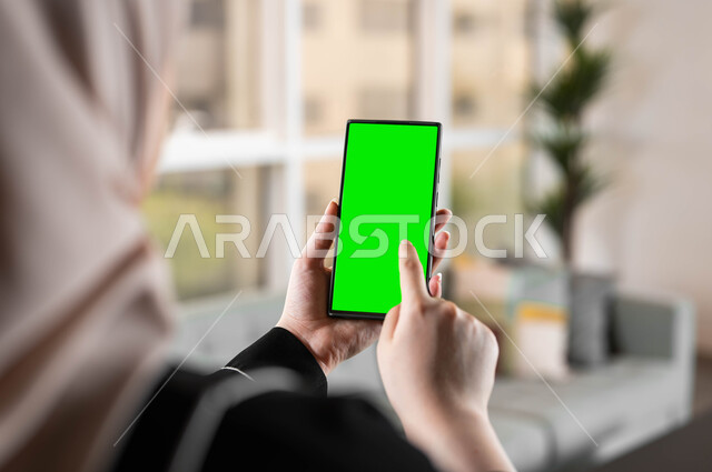 متابعة أخبار السوشال ميديا ومواقع التواصل الإجتماعي، استخدام تقنية سمارت على الهاتف المحمول للتحكم بالتلفاز، تصوير مقرب من الخلف لامرأة عربية خليجية سعودية تحمل بيدها موبايل بشاشة خضراء فارغة، الدردشات والمحادثات مع العائلة والأصدقاء، كروما