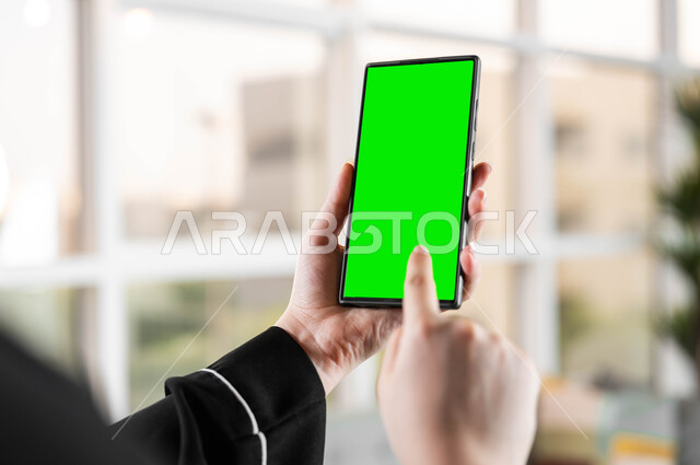 استخدام تقنية سمارت على الهاتف المحمول للتحكم بالتلفاز، تصوير مقرب من الخلف لامرأة عربية خليجية سعودية تحمل بيدها موبايل بشاشة خضراء فارغة، متابعة أخبار السوشال ميديا ومواقع التواصل الإجتماعي، الدردشات والمحادثات مع العائلة والأصدقاء، كروما