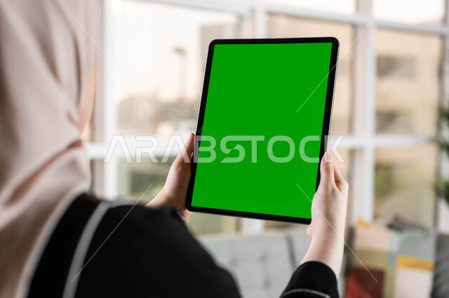 متابعة منصات التعليم الإلكتروني عبر الإنترنت، صورة مقربة من الخلف لامرأة عربية خليجية سعودية تقوم بحمل جهاز لوحي بشاشة خضراء فارغة، استخدام التكنولوجيا المتطورة والأجهزة التقنية الحديثة، إجراء مكالمات الفيديو مع الأصدقاء والأقارب، كروما