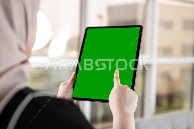 إجراء مكالمات الفيديو مع الأصدقاء والأقارب، صورة مقربة من الخلف لامرأة عربية خليجية سعودية تقوم بالنقر على جهاز لوحي بشاشة خضراء فارغة، استخدام التكنولوجيا المتطورة والأجهزة التقنية الحديثة، متابعة منصات التعليم الإلكتروني عبر الإنترنت، كروما