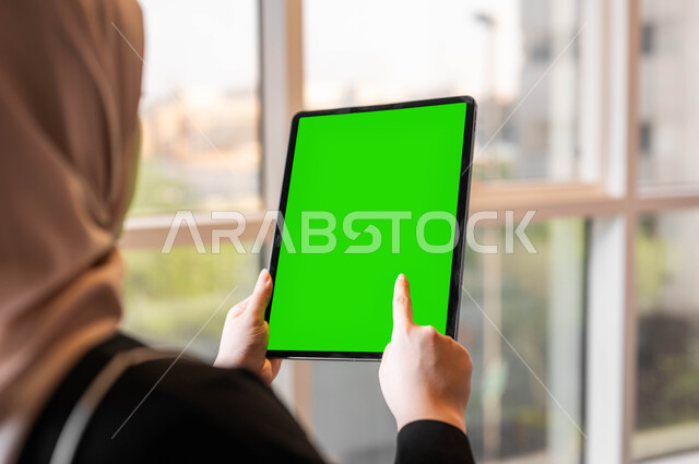 استخدام التكنولوجيا المتطورة والأجهزة التقنية الحديثة، متابعة منصات التعليم الإلكتروني عبر الإنترنت، صورة مقربة من الخلف لامرأة عربية خليجية سعودية تقوم بالنقر على جهاز لوحي بشاشة خضراء فارغة، إجراء مكالمات الفيديو مع الأصدقاء والأقارب، كروما