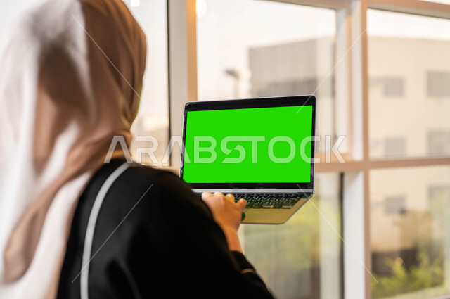 متابعة أخبار وسائل التواصل الإجتماعي، التطورات التقنية والتكنولوجية الحديثة، صورة مقربة من الخلف لامرأة عربية خليجية سعودية تحمل بيدها جهاز الحاسوب المحمول تقوم بمهام العمل عبر الإنترنت، أجهزة إلكترونية بشاشة خضراء فارغة، كروما