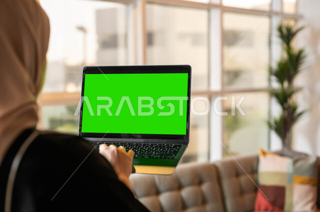 القيام بمهام العمل عبر الإنترنت، التطورات التقنية والتكنولوجية الحديثة، صورة مقربة من الخلف لامرأة عربية خليجية سعودية تحمل بيدها جهاز الحاسوب المحمول تقوم بمتابعة أخبار وسائل التواصل الإجتماعي، أجهزة إلكترونية بشاشة خضراء فارغة، كروما