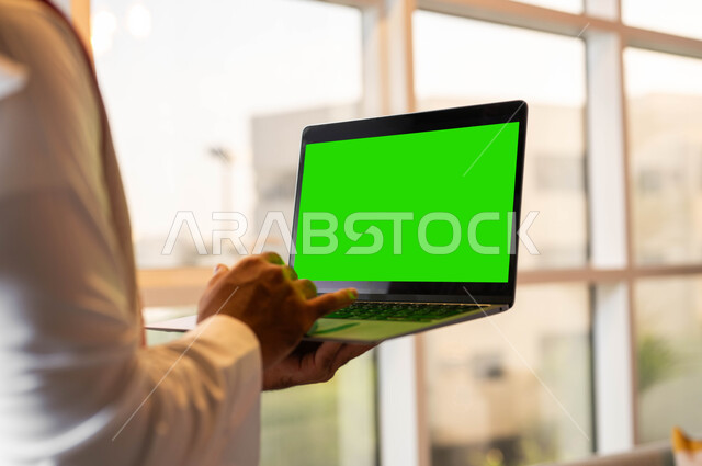 أجهزة إلكترونية بشاشة خضراء فارغة، إنجاز مهام العمل عبر الإنترنت، صورة مقربة من الخلف لرجل عربي خليجي سعودي يحمل بيده جهاز الحاسوب المحمول يقوم بمتابعة أخبار وسائل التواصل الإجتماعي، التطورات التقنية والتكنولوجية الحديثة، كروما