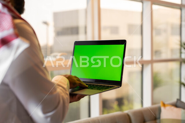 Completing work tasks online, a picture from behind of a Saudi Gulf Arab man holding a laptop in his hand, following social media news, electronic devices with a blank green screen, modern technical and technological developments, chroma