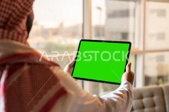 Technological and technical development in the fields of remote work, making video calls with friends and relatives, a picture from behind of a Saudi Gulf Arab man holding a tablet with a blank green screen in his hand, browsing social networking sites via the Internet, Chroma