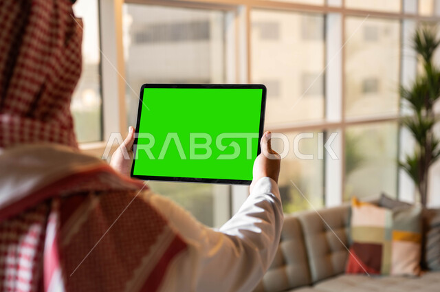 متابعة مواقع التواصل الإجتماعي عبر الإنترنت، إجراء مكالمات الفيديو مع الأصدقاء والأقارب، صورة مقربة من الخلف لرجل عربي خليجي سعودي يحمل بيده جهازالتابلت بشاشه خضراء فارغة، التطور التقني والتكنولوجي في مجالات العمل عن بعد، كروما