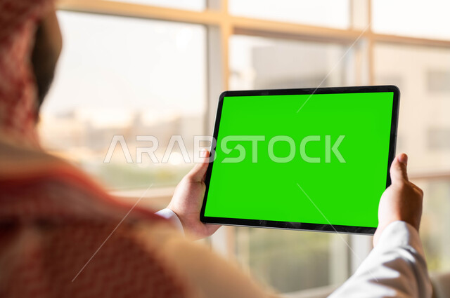 إجراء مكالمات الفيديو مع الأصدقاء والأقارب، تصفح مواقع التواصل الإجتماعي عبر الإنترنت، صورة مقربة من الخلف لرجل عربي خليجي سعودي يحمل بيده جهازالتابلت بشاشه خضراء فارغة، التطور التقني والتكنولوجي في مجالات العمل عن بعد، كروما