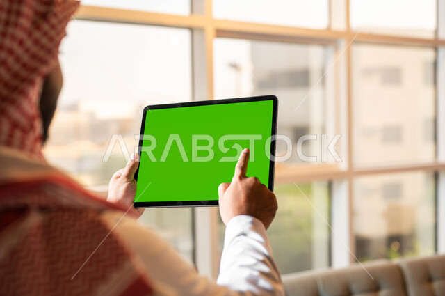 التطور التقني والتكنولوجي في مجالات العمل عن بعد، تصفح مواقع التواصل الإجتماعي عبر الإنترنت، صورة مقربة من الخلف لرجل عربي خليجي سعودي يقوم بالنقر على جهاز تابلت بشاشه خضراء فارغة، إجراء مكالمات الفيديو مع الأصدقاء والأقارب، كروما