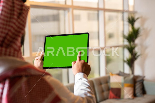 تصفح مواقع التواصل الإجتماعي عبر الإنترنت، صورة مقربة من الخلف لرجل عربي خليجي سعودي يقوم بالنقر على جهاز تابلت بشاشه خضراء فارغة، التطور التقني والتكنولوجي في مجالات العمل عن بعد، إجراء مكالمات الفيديو مع الأصدقاء والأقارب، كروما