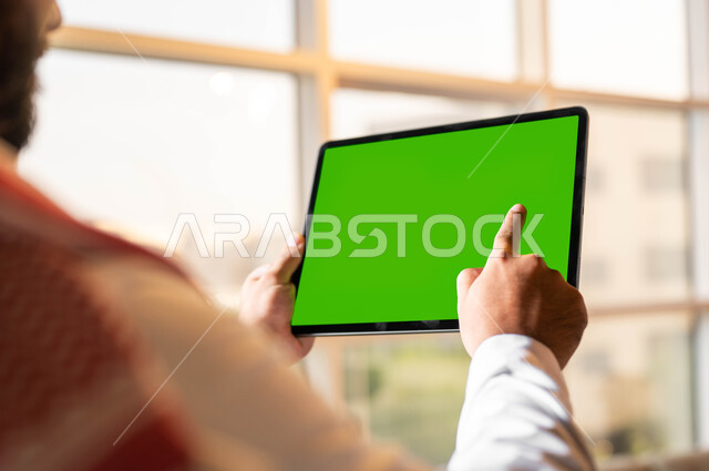 متابعة مواقع التواصل الإجتماعي عبر الإنترنت، صورة مقربة من الخلف لرجل عربي خليجي سعودي يرتدي الثوب التقليدي يحمل بيده جهاز التابليت بشاشه خضراء فارغة، التطور التكنولوجي في مجالات العمل عن بعد، دمج التقنية الحديثة بالحياة العملية, كروما