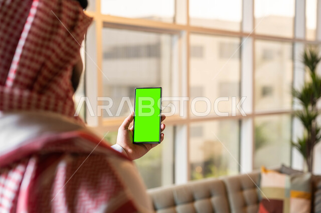 إجراء مكالمة فيديو ,التطور التكنولوجي و التقني في مجالات العمل عن بعد, إنجاز الأعمال باستخدام الهاتف المحمول، عرض شاشة خضراء فارغة على الأجهزة التقنية، صورة مقربة من الخلف لرجل عربي خليجي سعودي يرتدي الثوب التقليدي يحمل بيده الجوال، كروما
