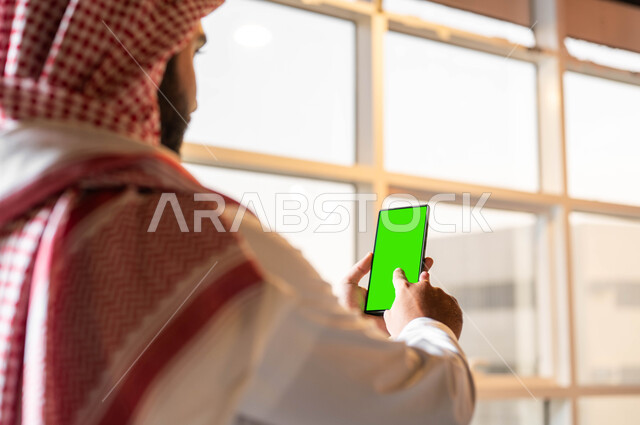 Technical and technological development in the fields of remote work, doing work using a mobile phone, a picture from the back of a Saudi Gulf Arab man wearing a traditional dress holding a mobile phone in his hand with a blank green screen, following the latest social media news, Chroma