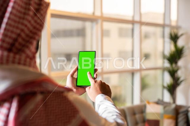 استخدام الأجهزة الإلكترونية الحديثة ,متابعة آخر أخبار السوشال ميديا، إنجاز مهام العمل عبر الإنترنت، صورة مقربة من الخلف لرجل عربي خليجي سعودي يرتدي الملابس التقليديه يحمل بيده الهاتف المحمول بشاشة خضراء فارغة، مفهوم اداره المشاريع عن بعد, كروما