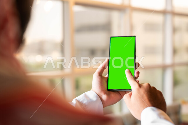Following the latest social media news, completing work tasks online, a close-up photo from the back of a Saudi Gulf Arab man wearing traditional clothing holding a mobile phone in his hand with a blank green screen, the concept of remote project management, using modern electronic devices, Chroma