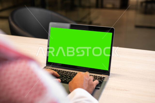 استعمال الحاسوب المحمول في إنجاز العمل، دمج التقنية والتكنولوجيا في الحياة العملية، متابعة مواقع التواصل الاجتماعي، عقد الاجتماعات عبر الانترنت، صورة مقربة من الخلف لرجل عربي خليجي سعودي يرتدي الزي التقليدي يقوم بالنقر على اللابتوب بشاشة خضراء فارغة، كروما