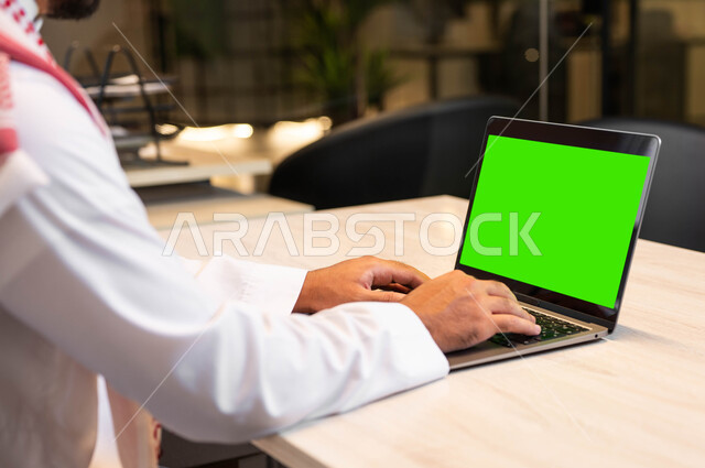 دمج التقنية والتكنولوجيا في الحياة العملية، متابعة مواقع التواصل الاجتماعي، عقد الاجتماعات عبر الانترنت، استعمال الحاسوب المحمول في إنجاز العمل، صورة مقربة من الخلف لرجل عربي خليجي سعودي يرتدي الزي التقليدي يقوم بالنقر على اللابتوب بشاشة خضراء فارغة، كروما