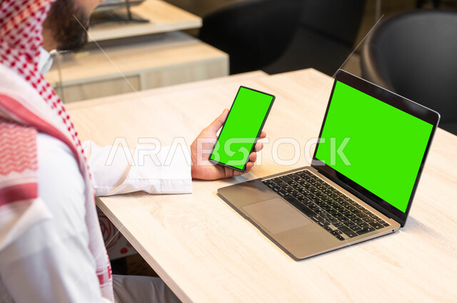 دمج التقنيات الحديثة في الحياة العملية، عرض شاشات خضراء فارغه عبر الاجهزة المتطورة ,اجراء اجتماعات العمل عبر الانترنت، صورة مقربة من الجانب لرجل عربي خليجي سعودي يرتدي الثوب التقليدي يحمل بيده الهاتف المحمول، استخدام اللابتوب في تنظيم خطط العمل، كروما