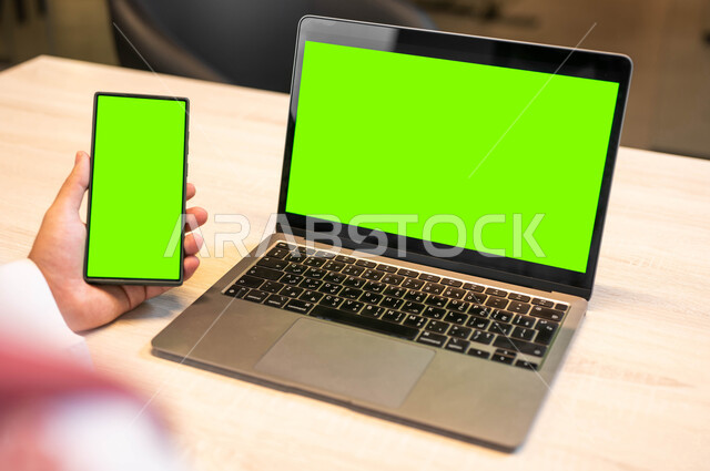 استخدام اللابتوب في تنظيم خطط العمل، عرض شاشات خضراء فارغه عبر الاجهزة المتطورة ,اجراء اجتماعات العمل عبر الانترنت، دور التقنية الحديثة في توفير راحة اكبر للافراد، صورة مقربة من الخلف لرجل عربي خليجي سعودي  يحمل بيده الهاتف المحمول، كروما