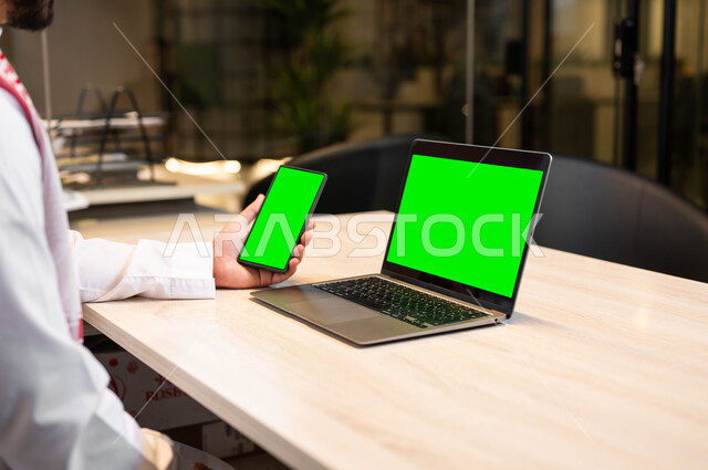 عرض شاشات خضراء فارغه عبر الاجهزة المتطورة ,اجراء اجتماعات العمل عبر الانترنت، دور التقنية الحديثة في توفير راحة اكبر للافراد، صورة مقربة من الجانب لرجل عربي خليجي سعودي يرتدي الثوب التقليدي يحمل بيده الهاتف المحمول، استخدام اللابتوب في تنظيم خطط العمل، كروما