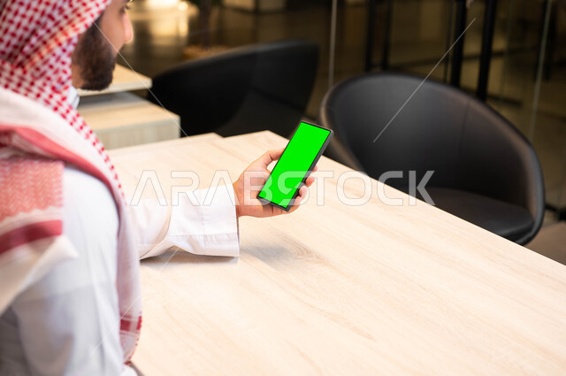 دور التقنية الحديثة في توفير راحة اكبر للافراد، صورة مقربة من الجانب لرجل عربي خليجي سعودي يرتدي الثوب التقليدي يحمل بيده الهاتف المحمول بشاشة خضراء فارغة، إنجاز مهام العمل عبر الإنترنت، التواصل مع الاهل و الاقارب، استخدام الأجهزة الإلكترونية ,كروما