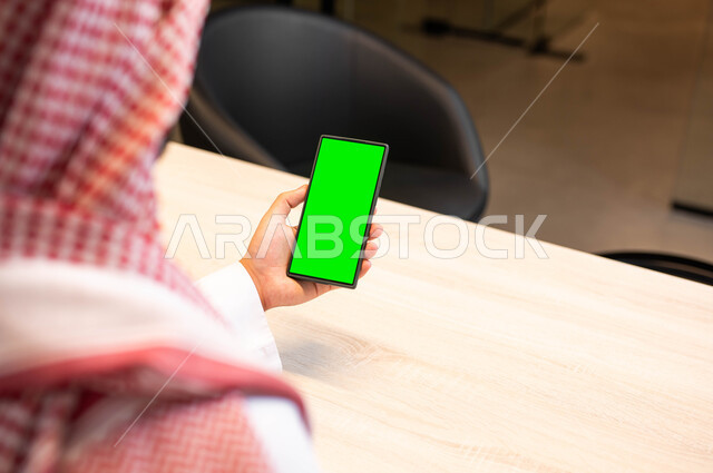 مشاهدة البرامج المفضلة في اوقات الفراغ, عرض شاشة خضراء فارغة على الأجهزة التقنية، استخدام التكنولوجيا الحديثة في انجاز المهام عن بعد، صورة مقربة من الخلف لرجل عربي خليجي سعودي يرتدي الزي التقليدي يحمل بيده الهاتف المحمول، كروما
