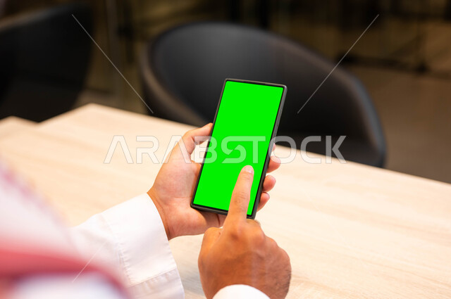 استخدام الأجهزة الحديثة الالكترونية, إنجاز مهام العمل عبر الإنترنت، متابعة مواقع التواصل الإجتماعي، صورة مقربة من الخلف لرجل عربي خليجي سعودي يرتدي الملابس التقليديه يحمل بيده الهاتف المحمول بشاشة خضراء فارغة، مفهوم اداره المشاريع عن بعد، كروما