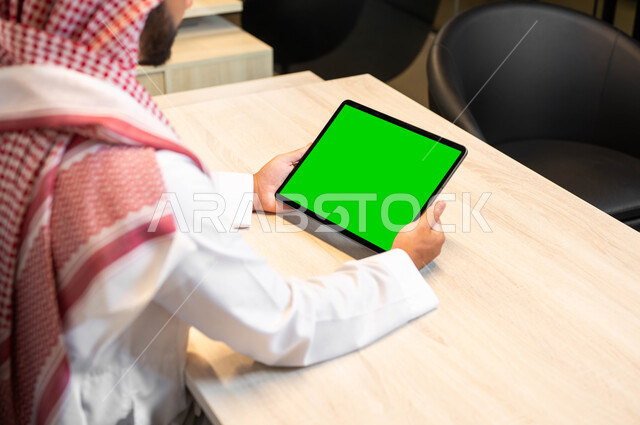 متابعة البرامج المفضلة عبر الانترنت, الجودة العالية للصوت والصورة على الأجهزة الحديثة، صورة مقربة من الخلف لرجل عربي خليجي سعودي يرتدي الزي التقليدي يحمل بيده جهاز التابليت بشاشة خضراء فارغه، استعمال التكنولوجيا في انجاز المهام عن بعد، كروما