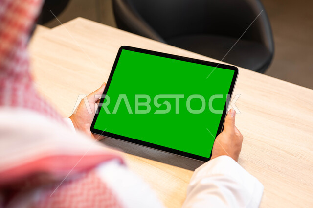الجودة العالية للصوت والصورة على الأجهزة الحديثة، صورة مقربة من الخلف لرجل عربي خليجي سعودي يرتدي الزي التقليدي يحمل بيده جهاز التابليت بشاشة خضراء فارغه، استعمال التكنولوجيا في انجاز المهام عن بعد، متابعة البرامج المفضلة عبر الانترنت, كروما