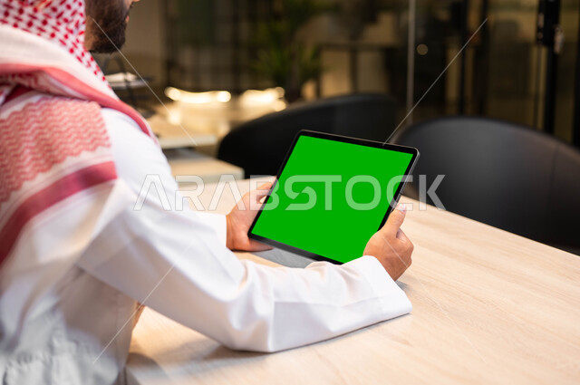 Using technology to complete tasks remotely, high-quality sound and image on modern devices, a close-up photo from the back of a Saudi Gulf Arab man wearing traditional clothing holding a tablet in his hand with a blank green screen, watching favorite programs online, Chroma