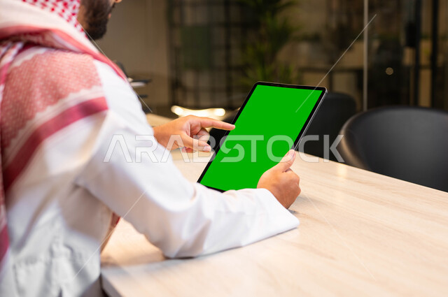 التواصل و الدردشة مع الاقارب, القيام بمقابلات وإجتماعات العمل أونلاين، استخدام الأجهزة التقنية الحديثة لتصفح الصور والفيديوهات بسهولة، صورة مقربة من الخلف لرجل عربي خليجي سعودي يرتدي الزي التقليدي يحمل بيده جهاز التابلت بشاشه خضراء فارغة، كروما