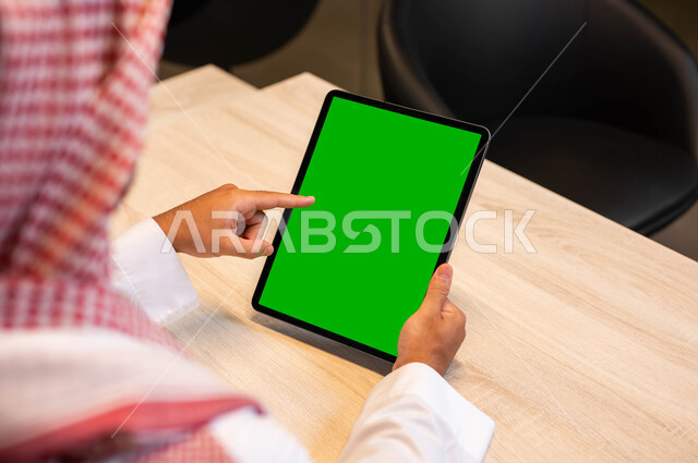 Conducting interviews and work meetings online, using modern technical devices to easily browse photos and videos, a close-up photo from the back of a Saudi Gulf Arab man wearing traditional clothing holding a tablet in his hand with a blank green screen, communicating and chatting with relatives, Chroma