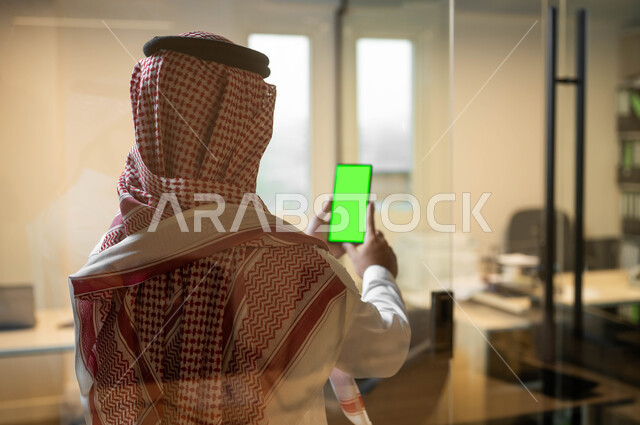 Technical and technological development in the fields of remote work, doing work using a mobile phone, using modern technical devices, a close-up photo from the back of a Saudi Gulf Arab man wearing a traditional thobe, displaying a blank green screen on a mobile phone, chroma
