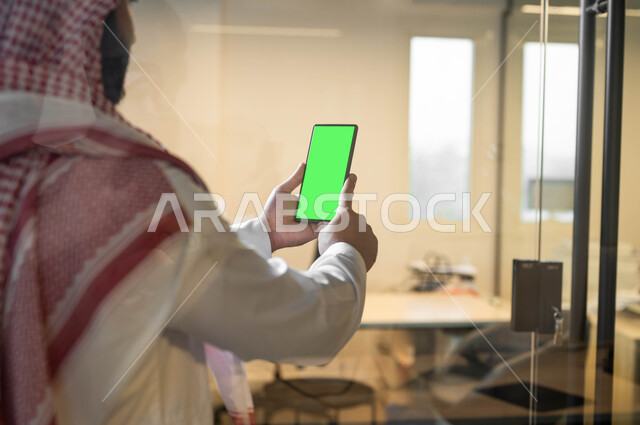 إجراء مكالمة فيديو عبر الجوال, استخدام الأجهزة التقنية الحديثة، اداره المشاريع عن بعد، صورة مقربة من الخلف لرجل عربي خليجي سعودي يرتدي الملابس التقليديه يحمل بيده الهاتف المحمول بشاشة خضراء فارغة، كروما
