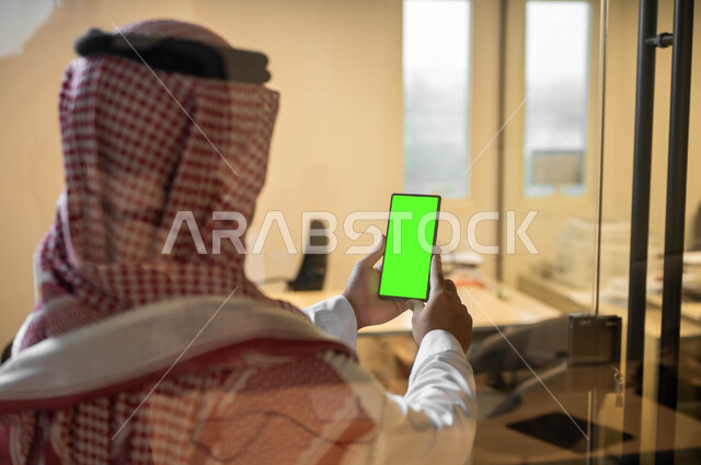 Holding meetings via the Internet, displaying a blank green screen on technical devices, a close-up picture from the back of a Saudi Gulf Arab man wearing a traditional dress holding a mobile phone in his hand, using modern technology to complete tasks remotely, Chroma