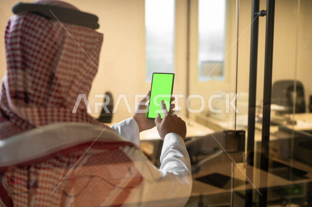 إنجاز مهام العمل عبر الإنترنت، متابعة مواقع التواصل الإجتماعي، صورة مقربة من الخلف لرجل عربي خليجي سعودي يرتدي الملابس التقليديه يحمل بيده الهاتف المحمول بشاشة خضراء فارغة، مفهوم اداره المشاريع عن بعد، استخدام الأجهزة الإلكترونية الحديثة, كروما