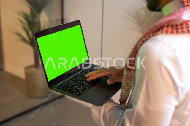 Completing work using a laptop, a close-up picture from the back of a Saudi Gulf Arab man wearing traditional clothing holding a laptop with a blank green screen in his hand, the concept of managing projects remotely, chatting and communicating with family, Chroma