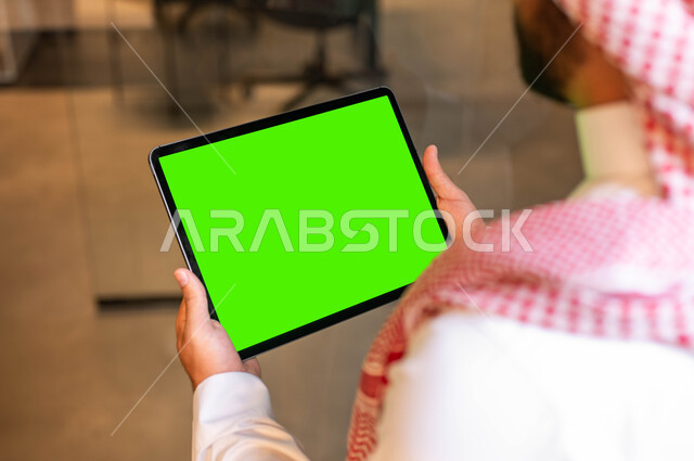 متابعة مواقع التواصل الإجتماعي عبر الإنترنت، صورة مقربة من الخلف لرجل عربي خليجي سعودي  يرتدي الزي التقليدي يحمل بيده جهاز التابلت بشاشه خضراء فارغة، إجراء مكالمات الفيديو مع الأصدقاء والأقارب، كروما