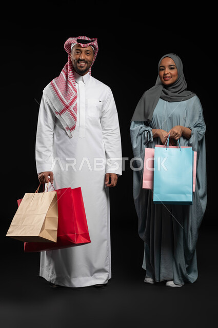 الاستفادة من التخفيضات والعروض الموسمية، مشاركة الزوجين في إختيار المشتريات، قضاء وقت ممتع بشراء الأغراض في الجمعة البيضاء، بورتريه لزوجان عربيان خليجيان سعوديان يحملان أكياس التسوق الملونة، تسوق المنتجات العالمية والسعودية، الطول الكامل للجسم، خلفية سوداء