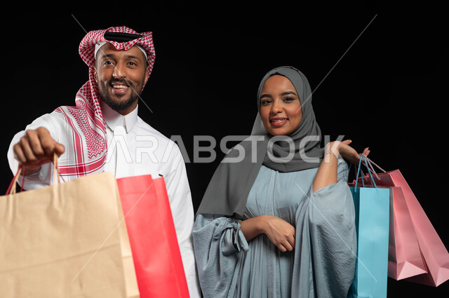 قضاء وقت ممتع بشراء الأغراض في الجمعة البيضاء، الاستفادة من التخفيضات والعروض الموسمية، مشاركة الزوجين في إختيار المشتريات، بورتريه مقرب لزوجان عربيان خليجيان سعوديان يحملان أكياس التسوق الملونة، تسوق المنتجات العالمية والسعودية، خلفية سوداء